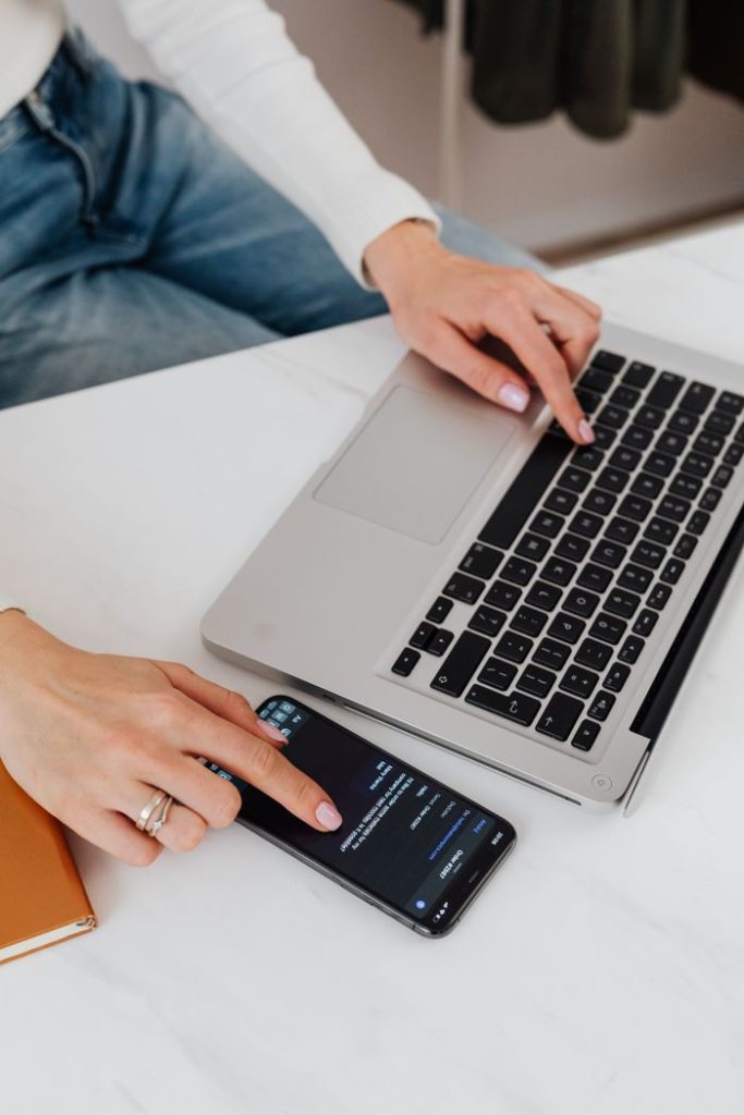 Woman multitasking with a laptop and smartphone on a desk in a well-lit office setting.